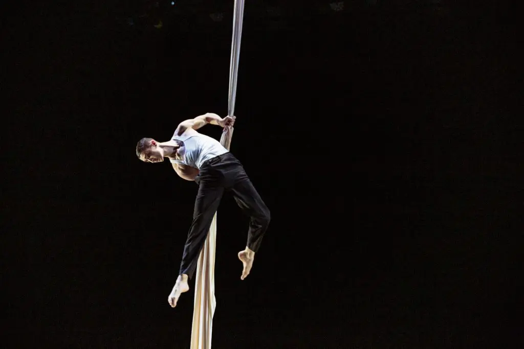 Antoine Boissereau, vêtu d'un débardeur blanc et d'un pantalon noir, effectue un numéro d'acrobatie aérienne en s'accrochant habilement à un tissu blanc suspendu sur un fond sombre.