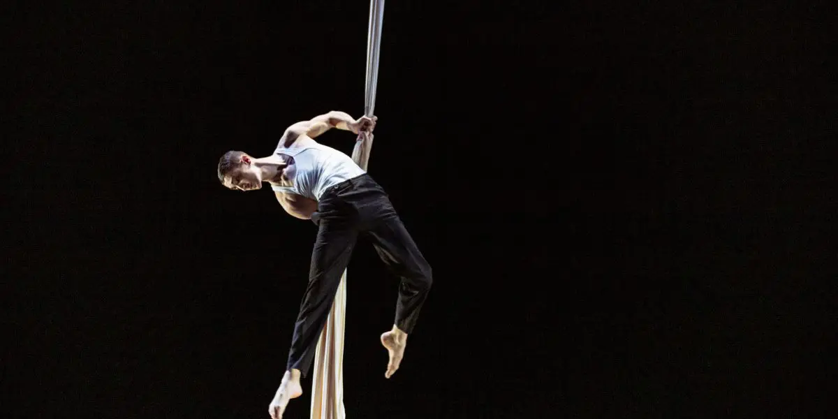 Antoine Boissereau, vêtu d'un débardeur blanc et d'un pantalon noir, effectue un numéro d'acrobatie aérienne en s'accrochant habilement à un tissu blanc suspendu sur un fond sombre.