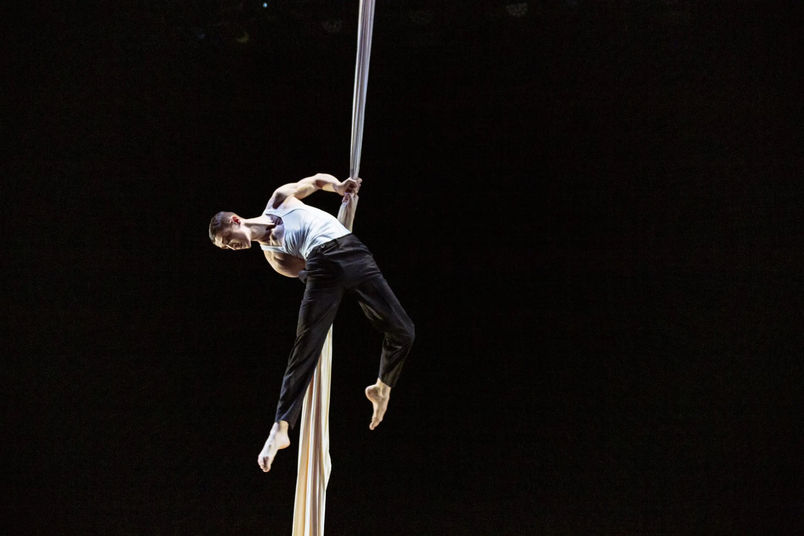 Antoine Boissereau, vêtu d'un débardeur blanc et d'un pantalon noir, effectue un numéro d'acrobatie aérienne en s'accrochant habilement à un tissu blanc suspendu sur un fond sombre.