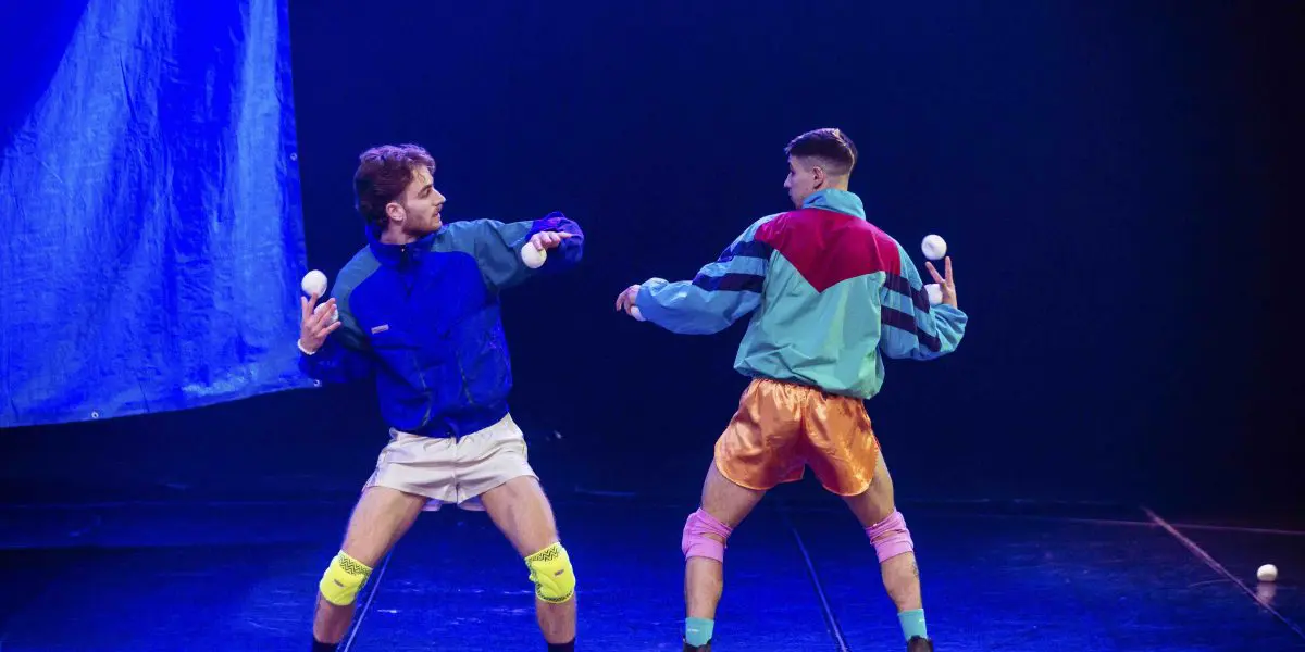 Deux artistes, Zachary Martel et Santiago Rivera Laugerud, vêtus de tenues rétro colorées, jonglent avec des balles blanches sur scène, prenant des poses dynamiques et se regardant dans les yeux sous un éclairage dramatique sur un fond bleu vif.