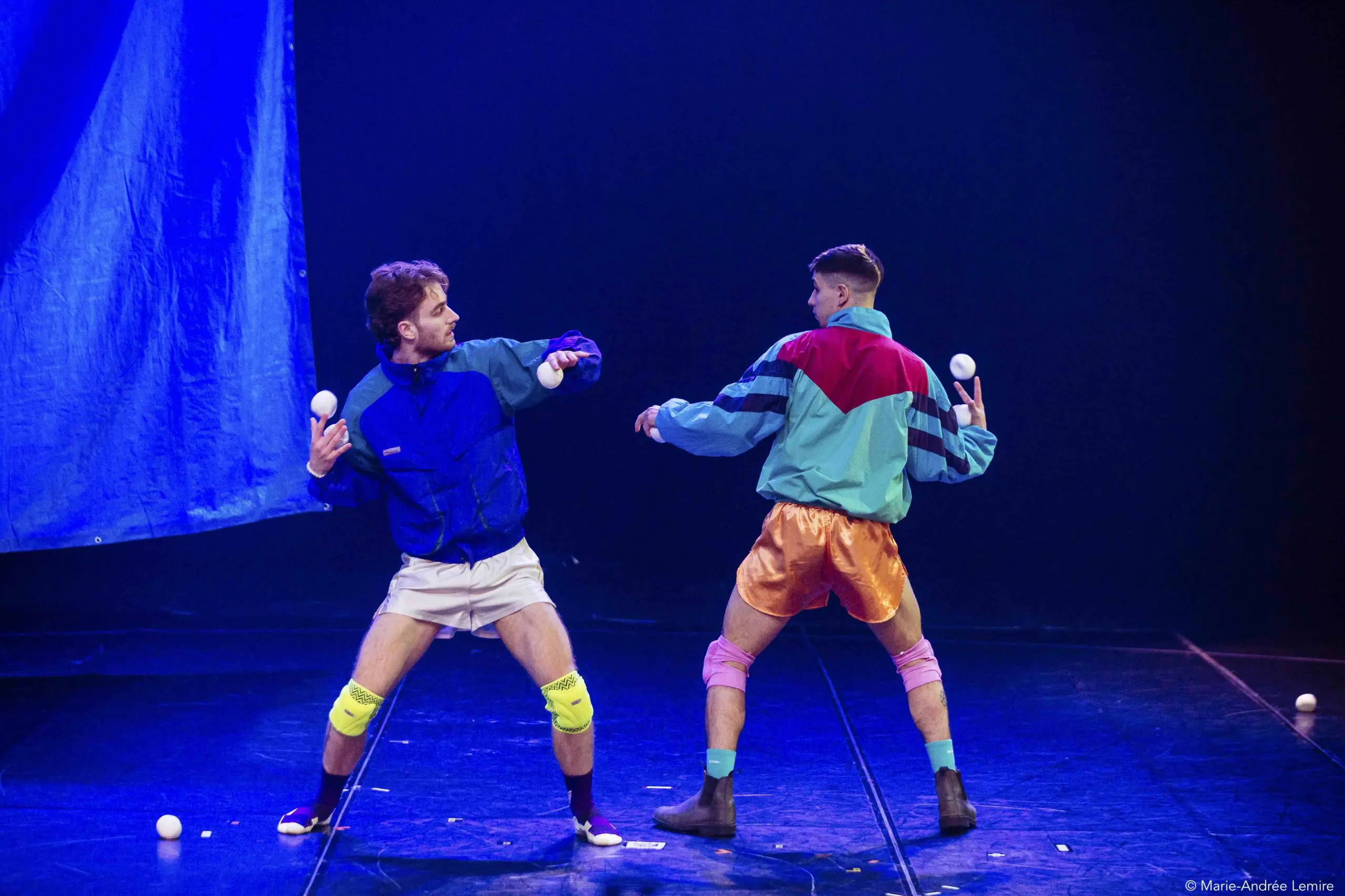 Deux artistes, Zachary Martel et Santiago Rivera Laugerud, vêtus de tenues rétro colorées, jonglent avec des balles blanches sur scène, prenant des poses dynamiques et se regardant dans les yeux sous un éclairage dramatique sur un fond bleu vif.