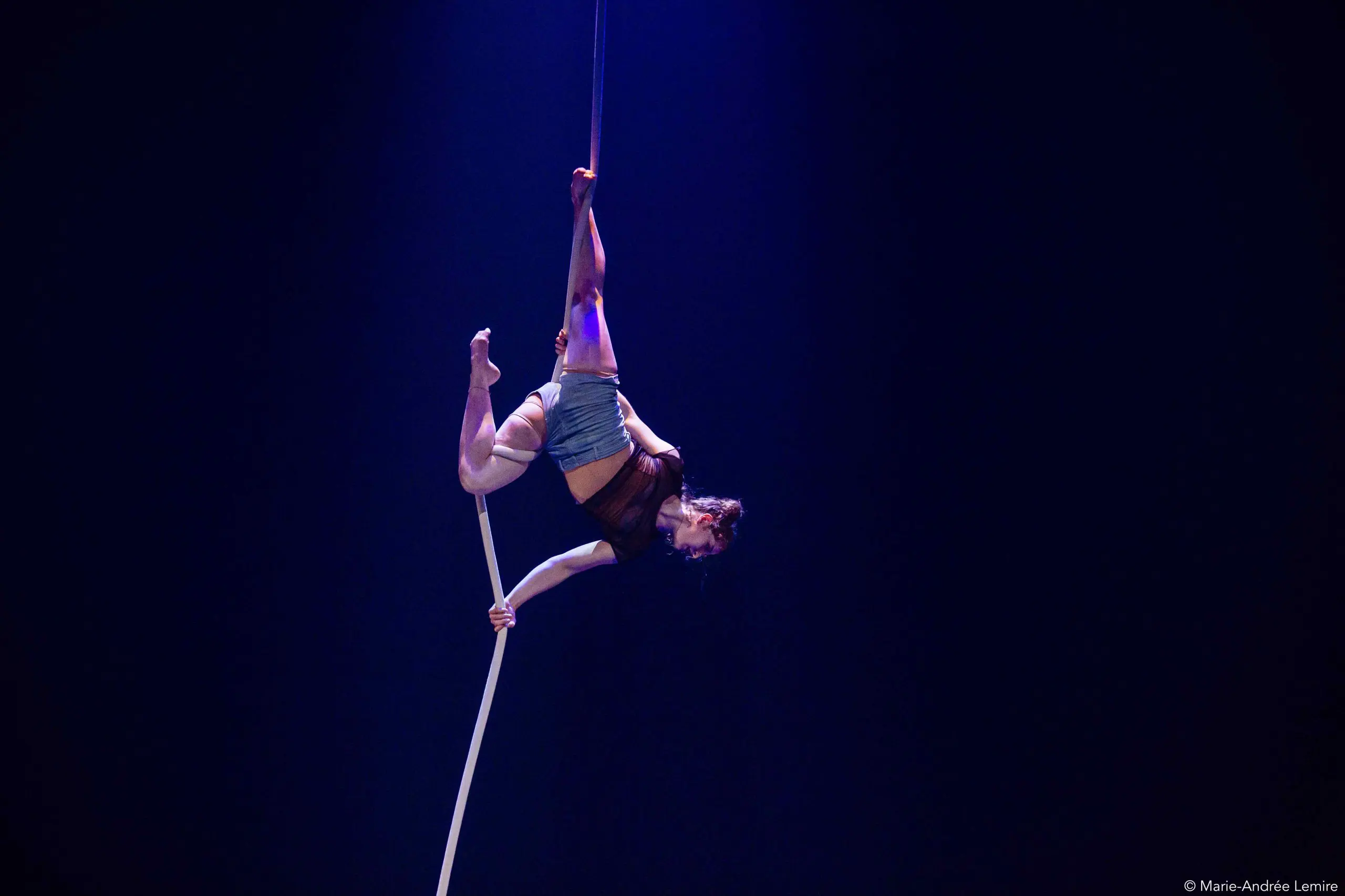 L'artiste de cirque Ellie Rossi exécute un numéro d'acrobatie aérienne, suspendue à l'envers par une jambe à une longue corde blanche, éclairée par un éclairage de scène dramatique sur un fond sombre.