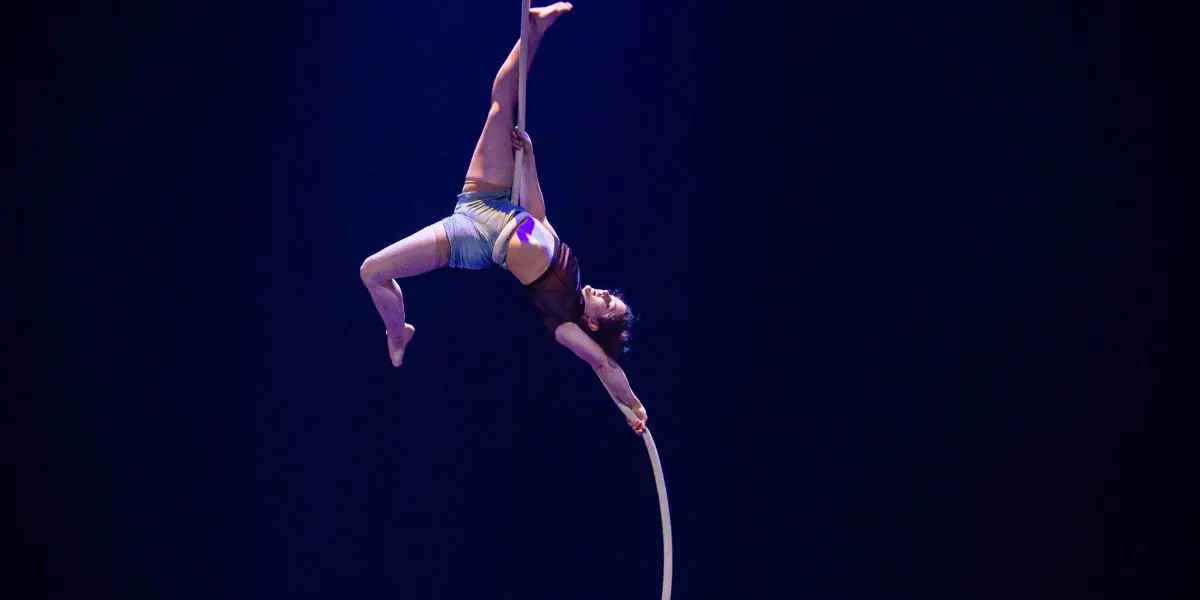 L'artiste de cirque Ellie Rossi, vêtue d'un haut et d'un short marron, est gracieusement suspendue la tête en bas à une corde aérienne blanche, une jambe et un bras tendus, sur un fond bleu foncé.
