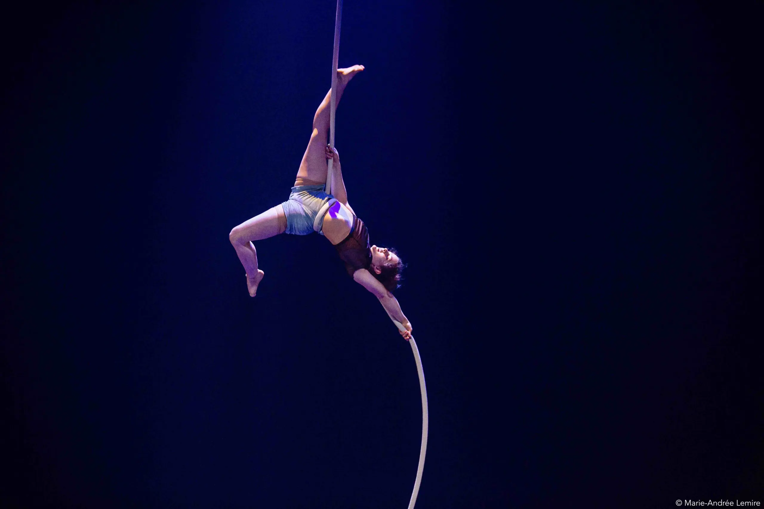 L'artiste de cirque Ellie Rossi, vêtue d'un haut et d'un short marron, est gracieusement suspendue la tête en bas à une corde aérienne blanche, une jambe et un bras tendus, sur un fond bleu foncé.