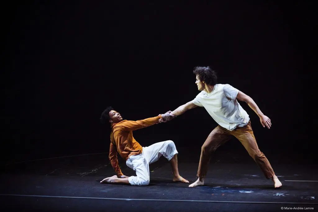 Deux danseurs évoluent sur une scène sombre ; Oliver Layher, vêtu d'une chemise blanche et d'un pantalon marron, se tient debout et tire l'autre, qui s'agenouille, vêtu d'un haut orange et d'un pantalon blanc - tous deux exprimant une émotion et un mouvement forts.