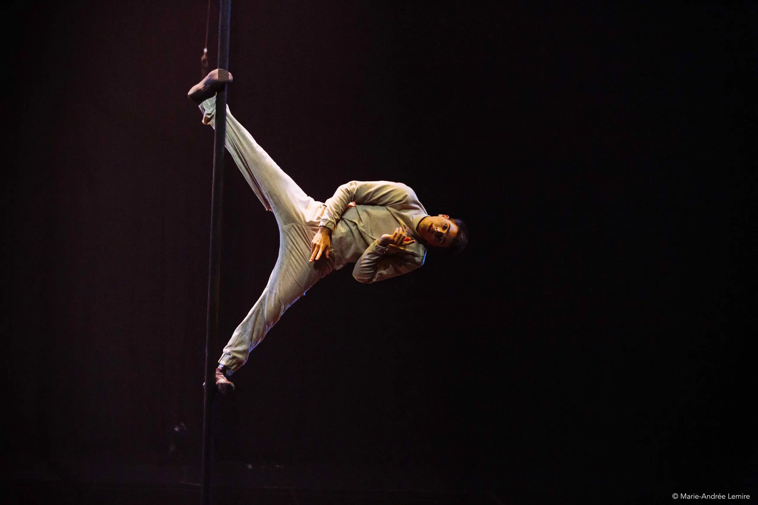 Un artiste vêtu d'une tenue gris clair se tient en équilibre horizontal sur un poteau vertical, s'agrippant d'un pied et d'une main, sur une scène sombre à l'éclairage dramatique.