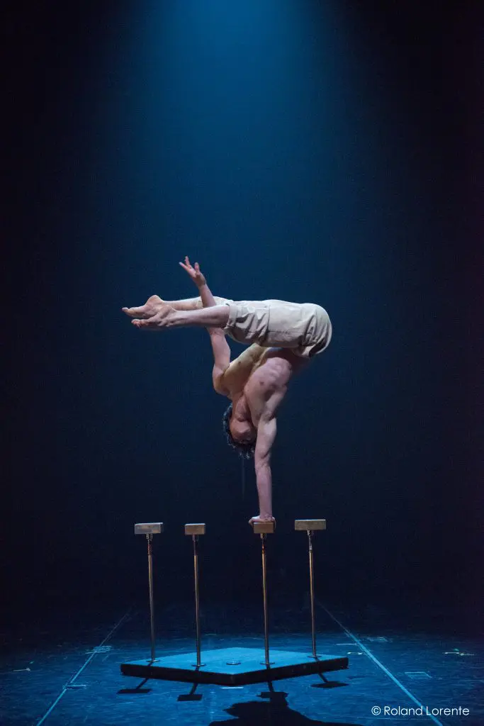 Un acrobate, Jarrod Takle, se tient en équilibre sur une main au sommet d'une paire de cannes sur une scène sombre, le corps arqué sur le côté, éclairé par un seul projecteur bleu.