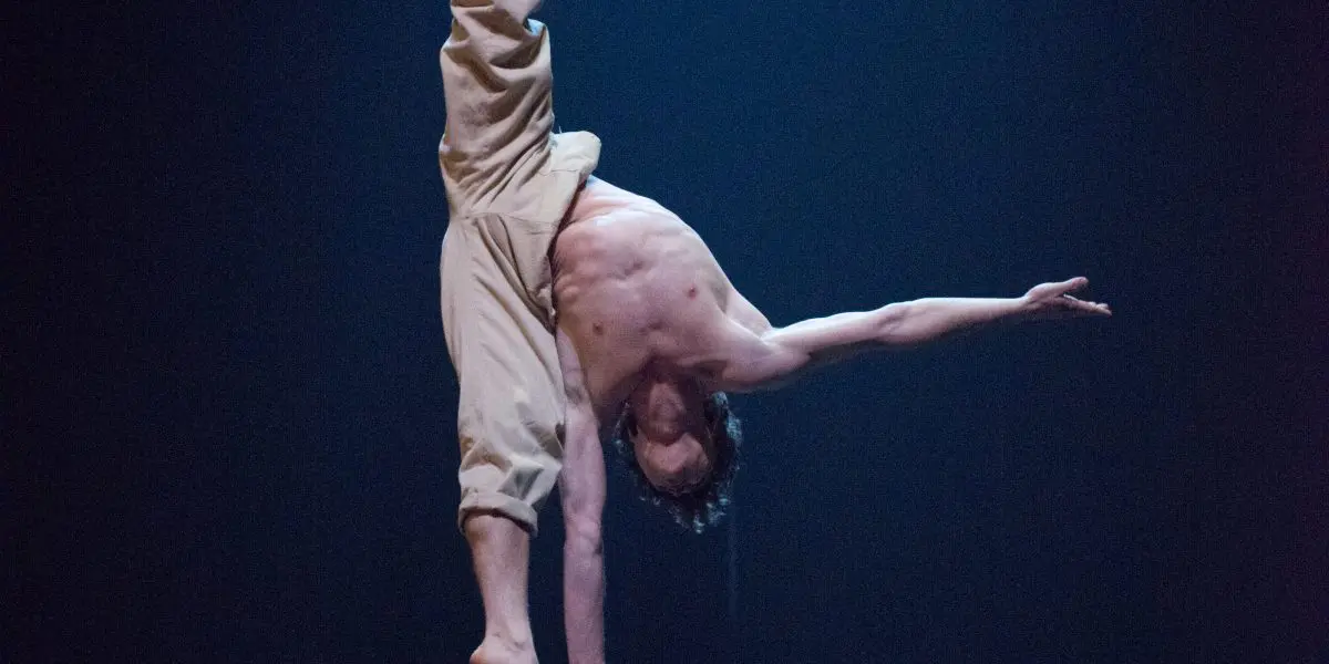 Jarrod Takle, torse nu, se tient en équilibre sur une main au sommet d'étroites plates-formes, les jambes écartées, sous un éclairage de scène dramatique et sur un fond sombre.