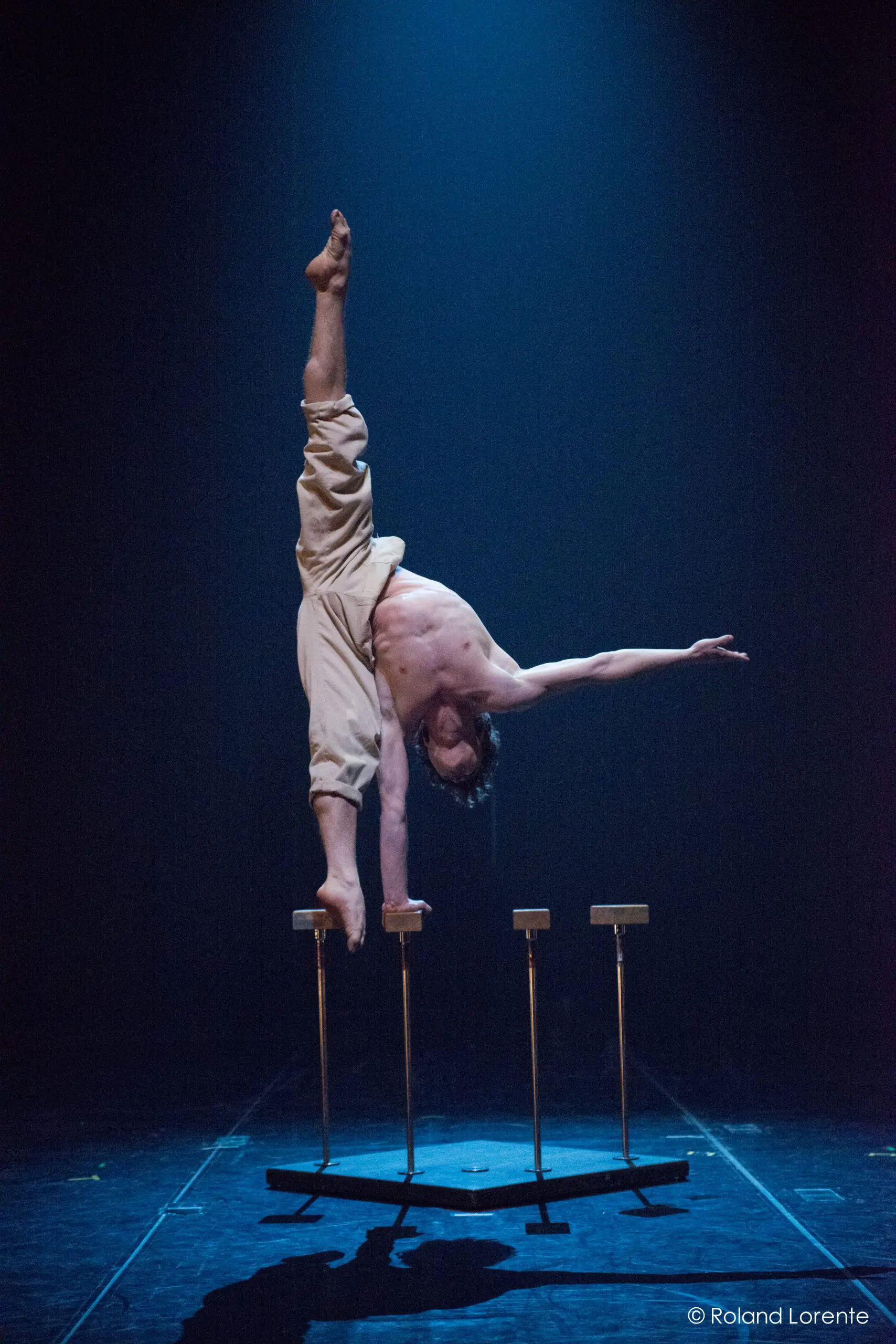 Jarrod Takle, torse nu, se tient en équilibre sur une main au sommet d'étroites plates-formes, les jambes écartées, sous un éclairage de scène dramatique et sur un fond sombre.
