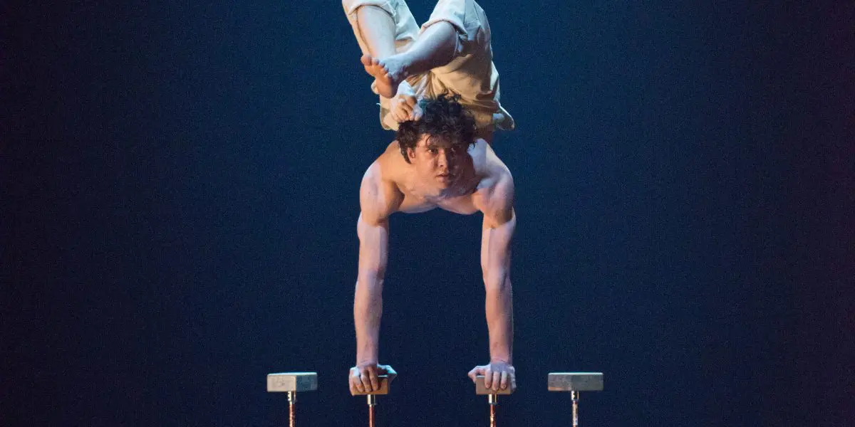 Un acrobate torse nu, Jarrod Takle, vêtu d'un pantalon blanc, effectue un poirier sur des barres parallèles sur scène, en arquant le dos et en repliant ses jambes au-dessus de sa tête sous un éclairage bleu spectaculaire.