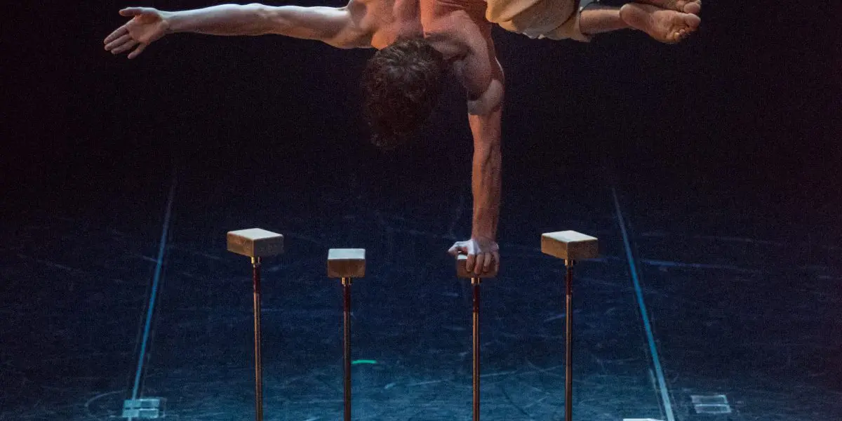 Jarrod Takle, un acrobate, se tient en équilibre horizontal sur une main au sommet d'un appareil à quatre supports sur une scène, les jambes superposées et un bras tendu vers l'extérieur. Un éclairage dramatique projette des ombres saisissantes sous lui.