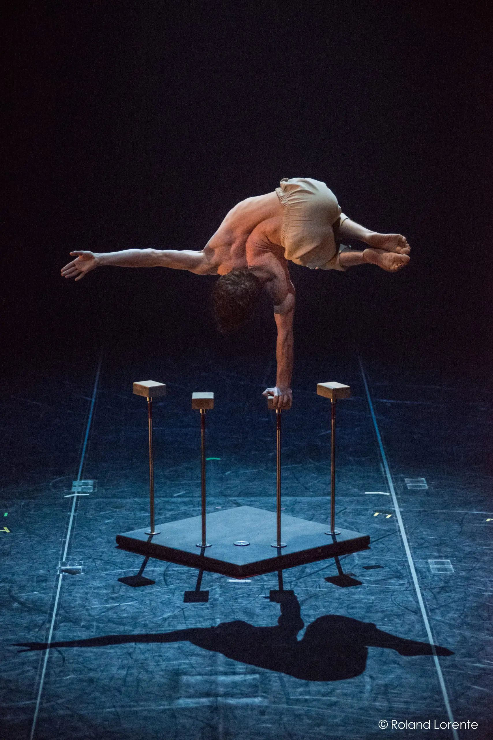 Jarrod Takle, un acrobate, se tient en équilibre horizontal sur une main au sommet d'un appareil à quatre supports sur une scène, les jambes superposées et un bras tendu vers l'extérieur. Un éclairage dramatique projette des ombres saisissantes sous lui.