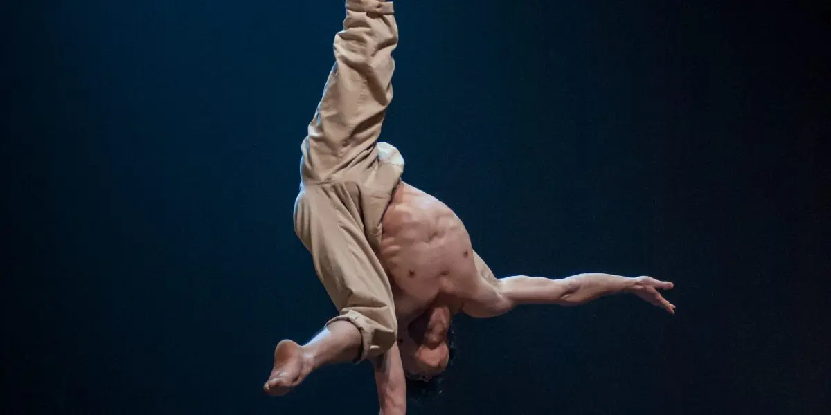 Jarrod Takle, torse nu, se tient en équilibre sur une main au sommet d'une paire de cannes surélevées, les jambes tendues vers le haut en position fendue, sur une scène sombre à l'éclairage dramatique.