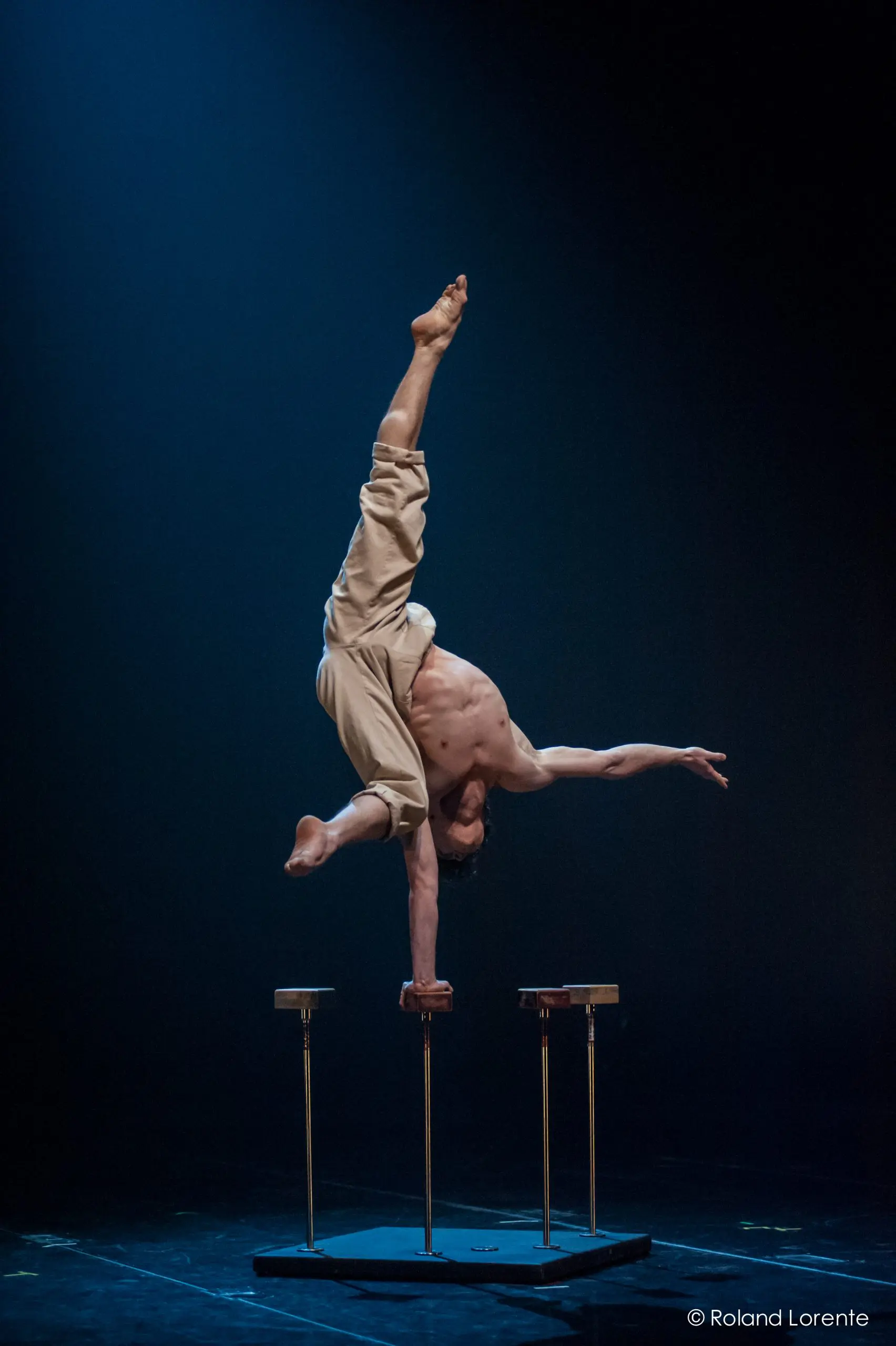 Jarrod Takle, torse nu, se tient en équilibre sur une main au sommet d'une paire de cannes surélevées, les jambes tendues vers le haut en position fendue, sur une scène sombre à l'éclairage dramatique.
