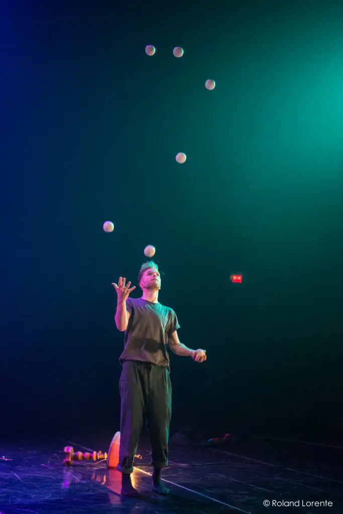 Une personne, peut-être John Witte, se tient sur scène sous un éclairage dramatique, jonglant avec sept balles blanches en arc de cercle au-dessus de sa tête. Des quilles de jonglage et des accessoires sont éparpillés sur le sol à proximité.