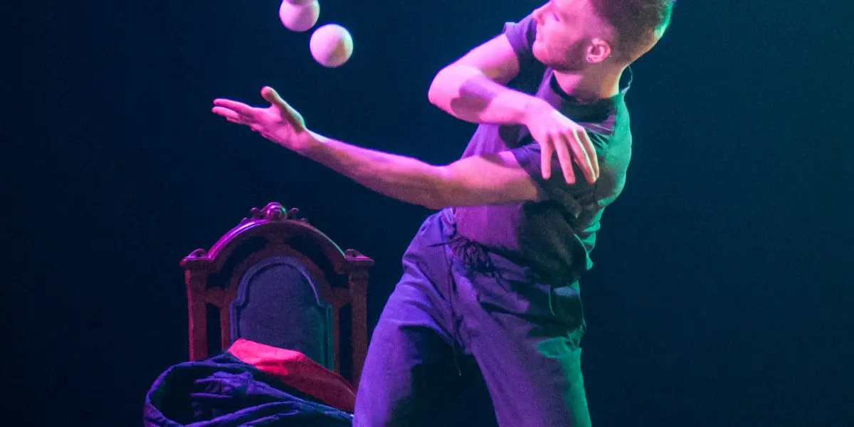 Un artiste vêtu de vêtements sombres jongle habilement avec quatre balles blanches sur scène, à côté d'une chaise antique, sous un éclairage dramatique - un art qui rappelle celui de John Witte.