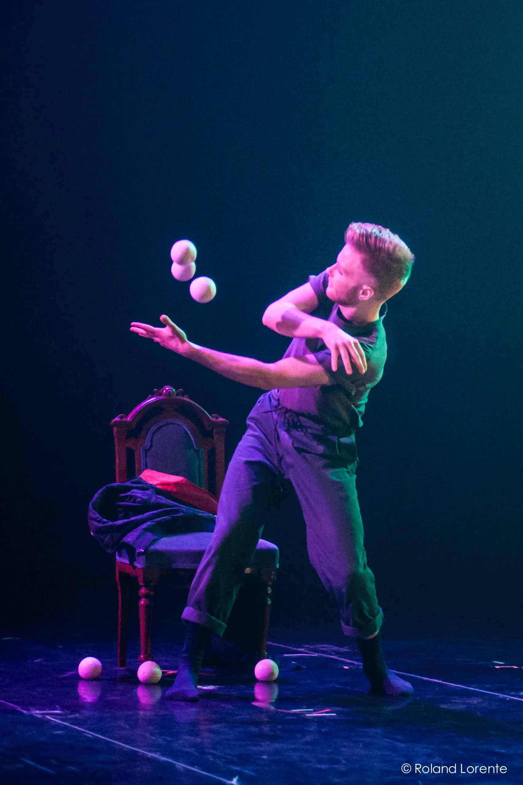 Un artiste vêtu de vêtements sombres jongle habilement avec quatre balles blanches sur scène, à côté d'une chaise antique, sous un éclairage dramatique - un art qui rappelle celui de John Witte.