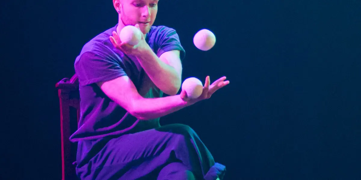 John Witte est assis sur une chaise sur scène, jonglant avec cinq balles blanches avec une grande concentration sous un éclairage dramatique et coloré.
