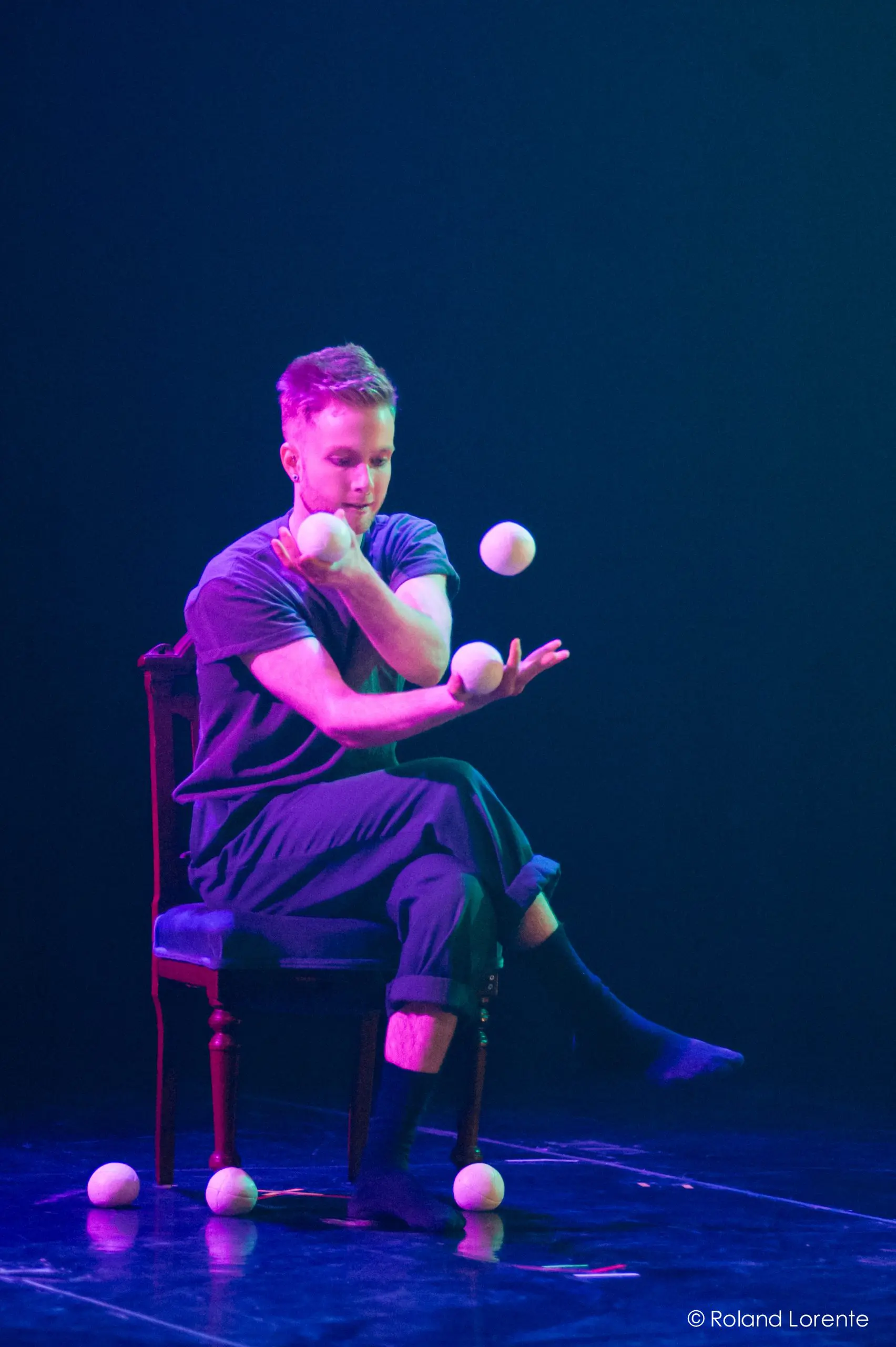 John Witte est assis sur une chaise sur scène, jonglant avec cinq balles blanches avec une grande concentration sous un éclairage dramatique et coloré.