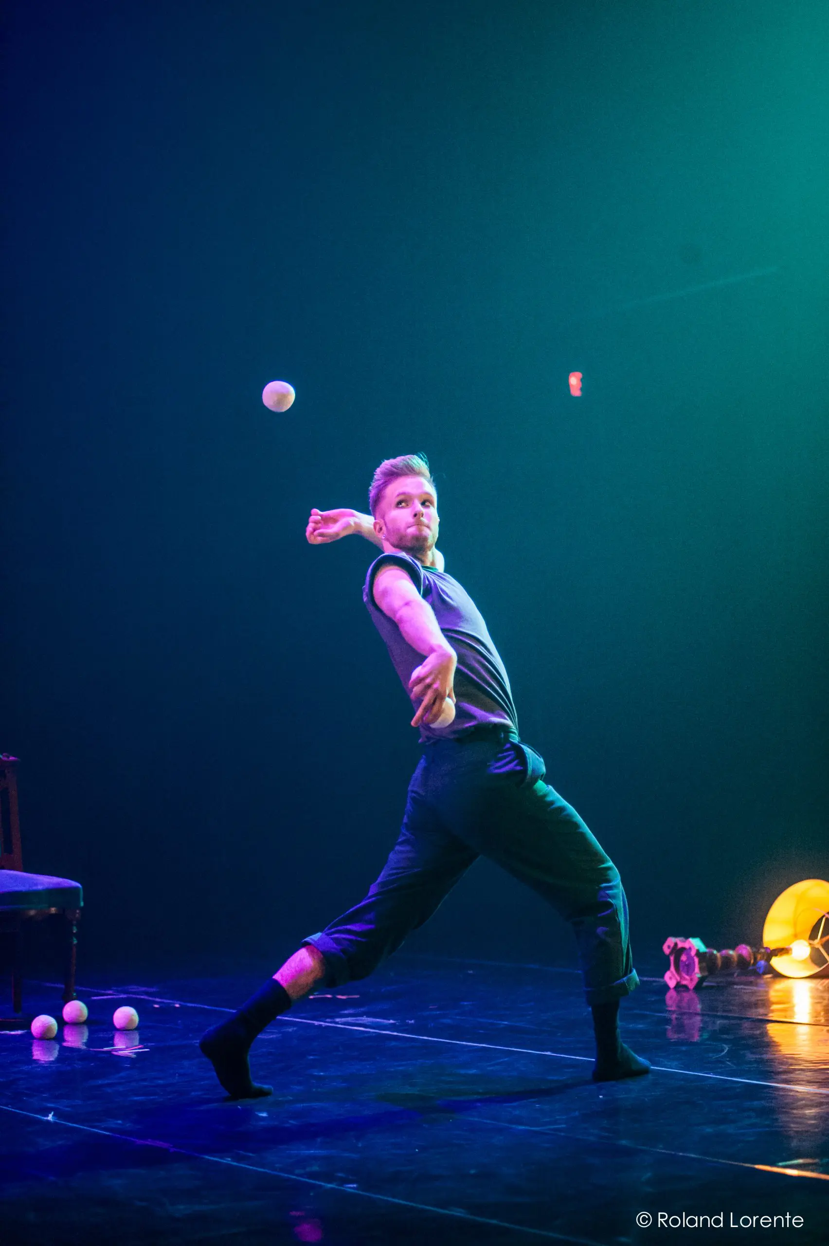 Un artiste, peut-être John Witte, jongle avec une balle sur scène dans un éclairage bleu et vert spectaculaire. Plusieurs balles reposent sur le sol près d'une chaise, et des jouets en peluche sont visibles à l'arrière-plan.