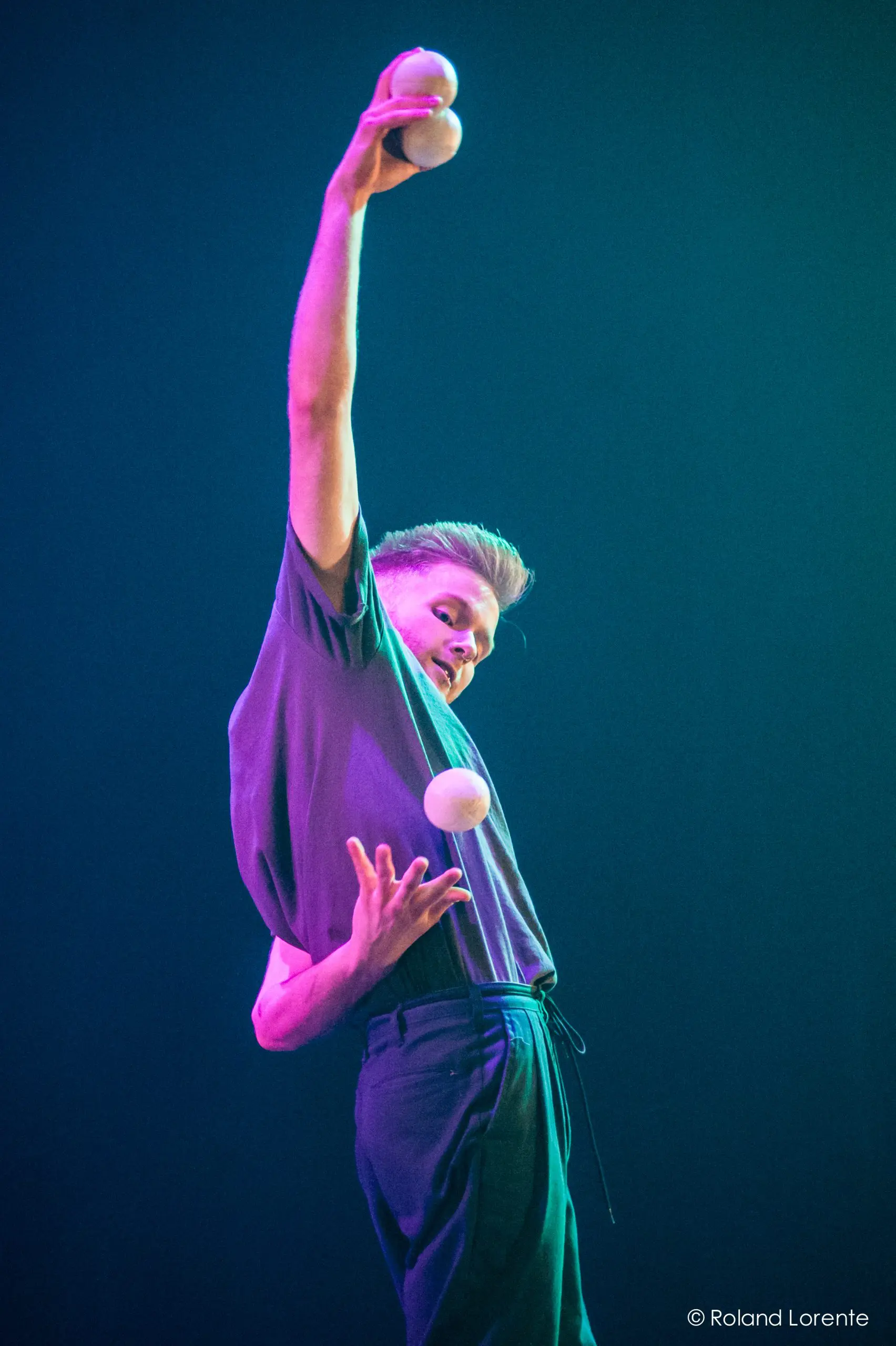 John Witte fait un numéro de jonglage sur scène, tenant en équilibre deux balles blanches dans une main et lançant une autre balle en l'air, avec un éclairage dramatique mettant en valeur son expression concentrée.
