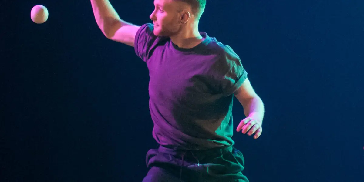 John Witte, vêtu d'un t-shirt et d'un pantalon sombres, jongle avec quatre balles sur une scène faiblement éclairée, un projecteur mettant en valeur son expression et ses mouvements concentrés.