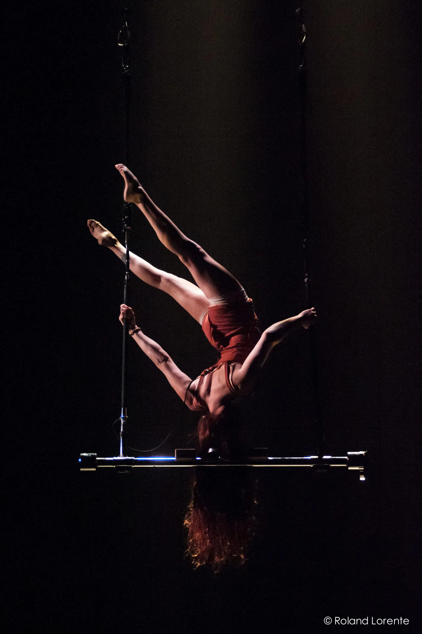 Une voltigeuse est suspendue à un trapèze, les bras et les jambes gracieusement tendus, sous les projecteurs, sur un fond sombre, illustrant l'art de Louana Seclet-Monchot.