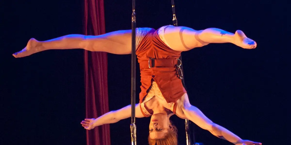Une voltigeuse, Louana Seclet-Monchot, vêtue d'un costume orange, exécute un grand écart à l'envers sur un trapèze, les bras étendus et les cheveux pendants, sur un fond de scène sombre.