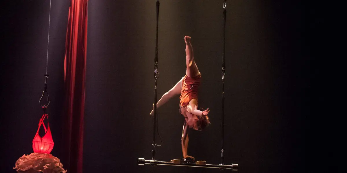 Une acrobate, Louana Seclet-Monchot, vêtue d'une tenue orange, se tient en équilibre sur une main sur un trapèze suspendu, en étendant ses jambes vers le haut. Un éclairage de scène doux la met en valeur sur un fond sombre, avec de la soie aérienne rouge et des tissus drapés à proximité.