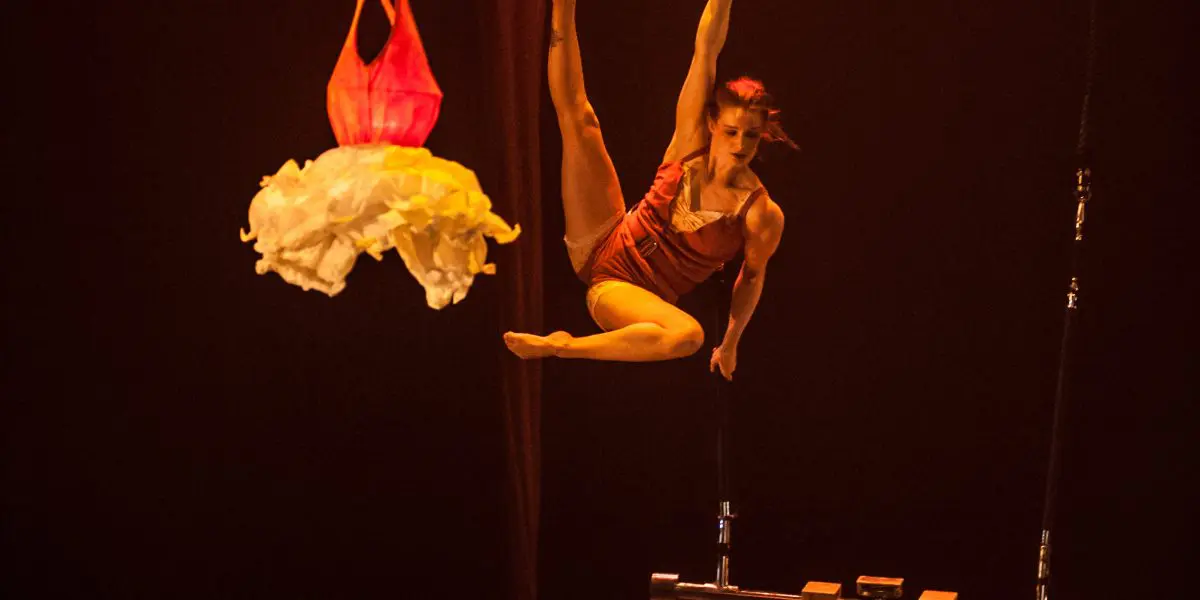 Une voltigeuse, Louana Seclet-Monchot, se produit sur scène, suspendue à un tissu rouge, une jambe tendue et les bras agrippés au tissu. Un costume blanc à volants est suspendu à proximité et un trapèze est visible dans l'atmosphère tamisée et chaudement éclairée.