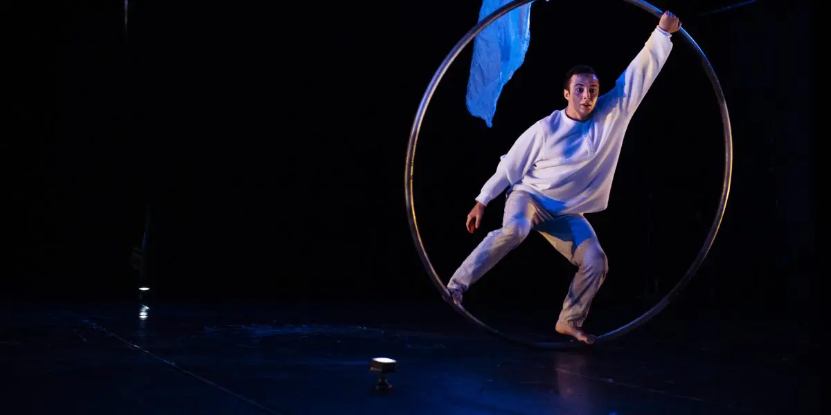 Un artiste vêtu de blanc se tient en équilibre à l'intérieur d'un grand cerceau métallique sur une scène sombre, avec un tissu bleu suspendu au-dessus et un éclairage dramatique mettant en valeur la scène - un numéro caractéristique de Robert Cookson.