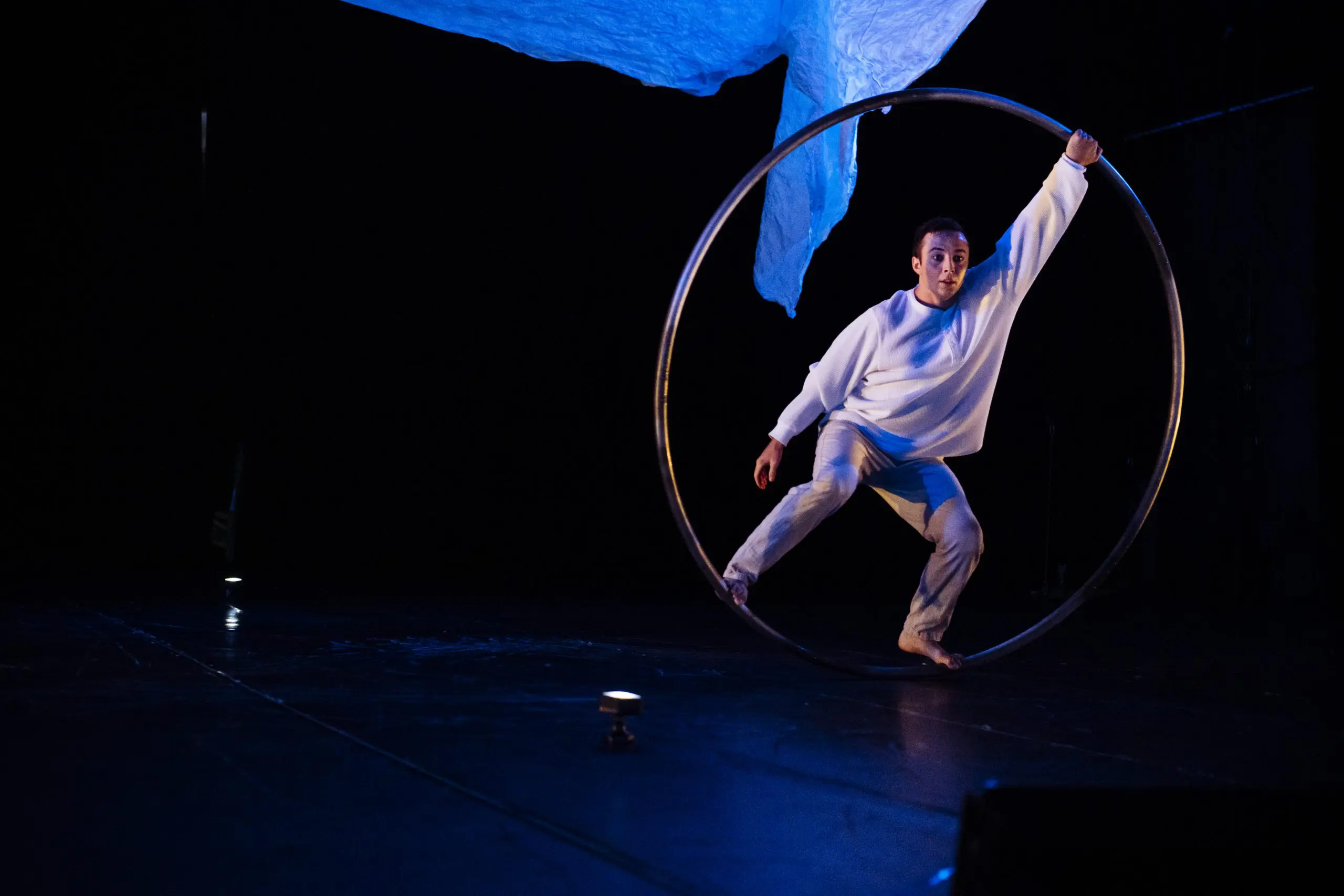 Un artiste vêtu de blanc se tient en équilibre à l'intérieur d'un grand cerceau métallique sur une scène sombre, avec un tissu bleu suspendu au-dessus et un éclairage dramatique mettant en valeur la scène - un numéro caractéristique de Robert Cookson.