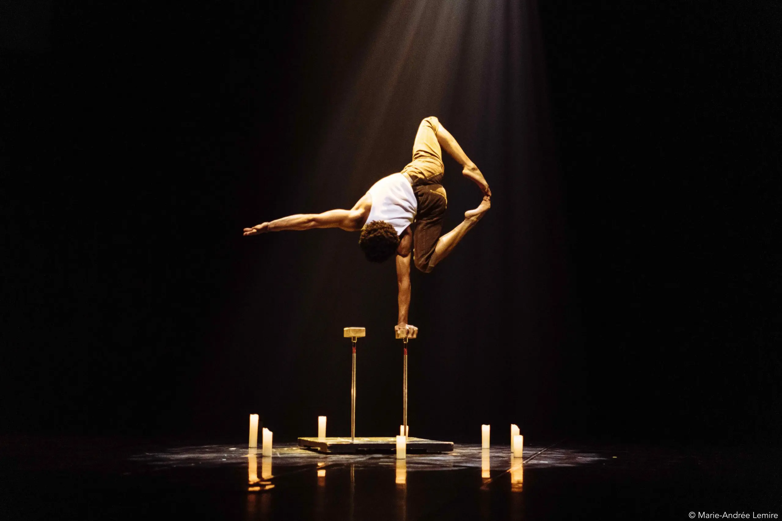Un acrobate, Kyran Lee Walton, se tient en équilibre sur une main au sommet de cannes, les jambes étendues en fente, éclairé par un projecteur sur une scène sombre entourée de bougies incandescentes.