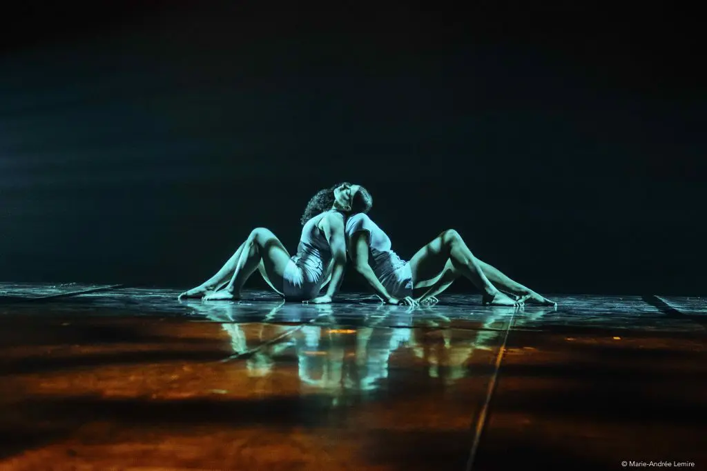 Deux danseuses, Léann Gingras et Kaisha Dessalines Wright, sont assises dos à dos sur une scène faiblement éclairée, les jambes étendues et les bras entrelacés, créant une pose symétrique en miroir sous un éclairage dramatique.