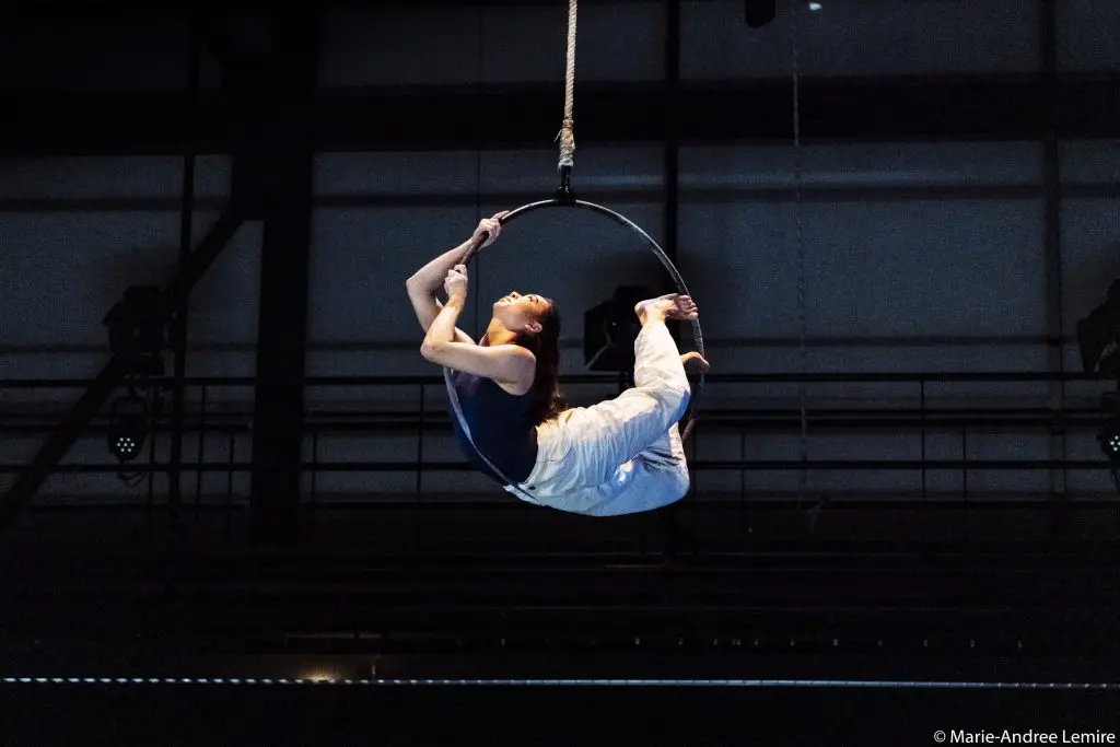 Michelle Sargent, une artiste de cirque vêtue d'un pantalon blanc et d'un haut noir, se tient en équilibre la tête en bas sur un cerceau aérien suspendu, tenant une jambe tout en arquant le dos sur la scène faiblement éclairée.