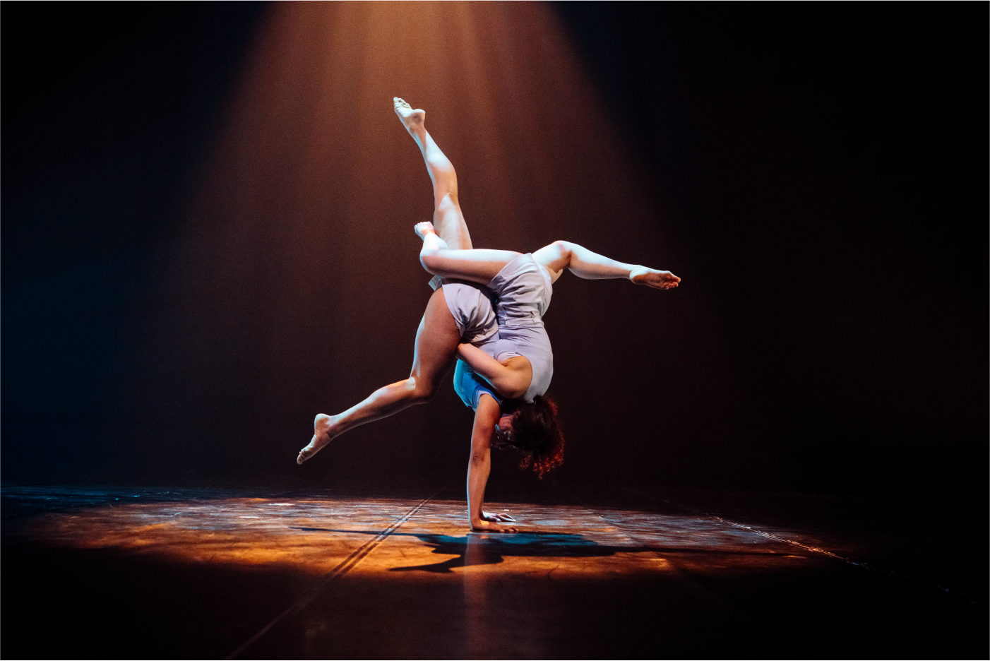 Une danseuse en tenue grise, rappelant l'élégance du conseil d'administration de l'ECN, se tient en équilibre sur une main, les jambes tendues en fente, effectuant un mouvement acrobatique sous les projecteurs d'une scène sombre.