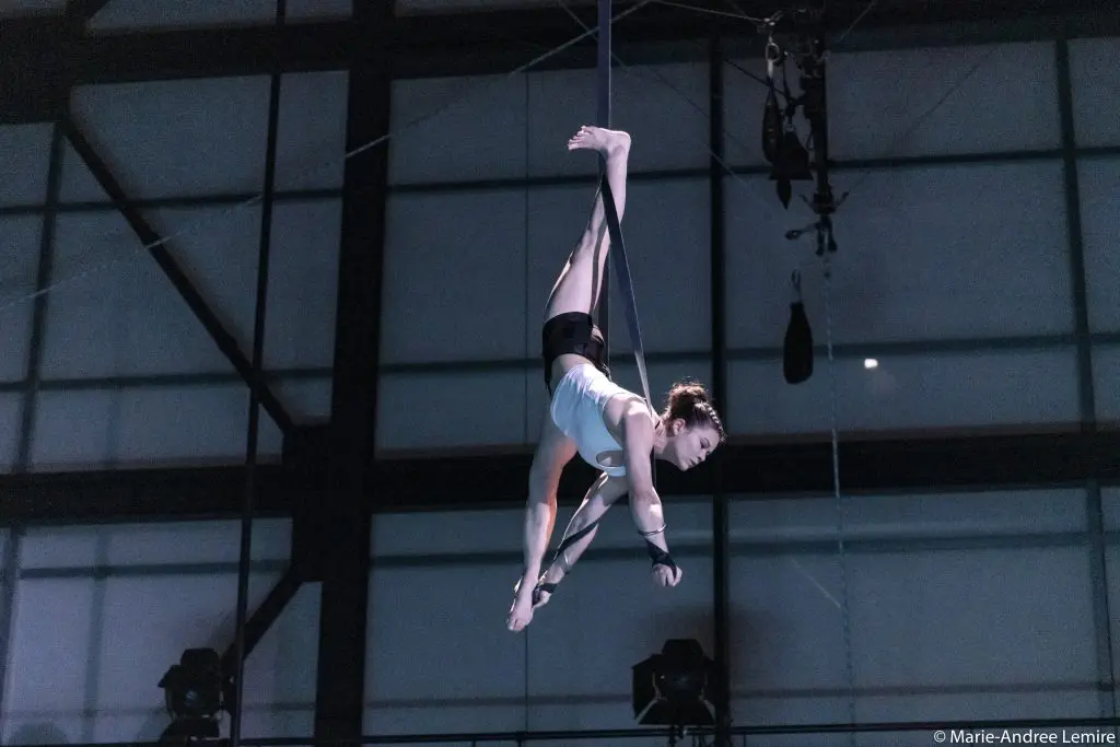 Clara Laurent, une voltigeuse vêtue d'un haut blanc et d'un short noir, effectue une pose à l'envers sur des sangles aériennes suspendues à l'intérieur d'un bâtiment de style industriel.