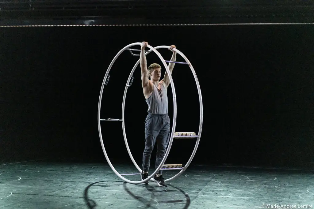 Sur une scène faiblement éclairée, Elijah Kirk se tient à l'intérieur d'une grande roue Cyr en métal, qu'il tient à deux mains. L'arrière-plan sombre met en valeur à la fois l'artiste et la silhouette frappante de la roue.