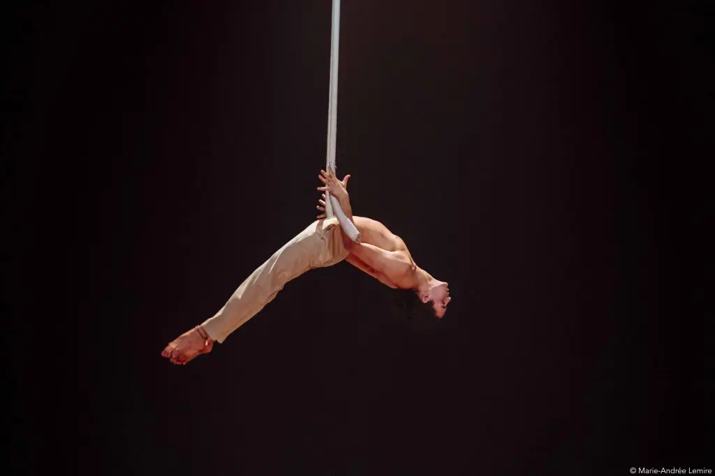 L'artiste aérien Zebulon Simoneau, vêtu d'un pantalon beige, est suspendu à l'envers à une corde, le dos arqué et les bras tendus, sur un fond sombre.