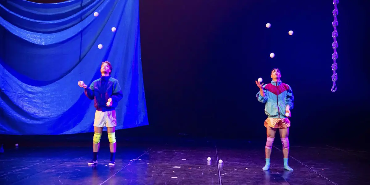 Deux jongleurs, Zachary Martel et Santiago Rivera Laugerud, se produisent sur scène, chacun lançant plusieurs balles blanches en l'air. Ils portent des tenues colorées et se tiennent sous un éclairage bleu spectaculaire, sur une toile de fond bleue drapée.