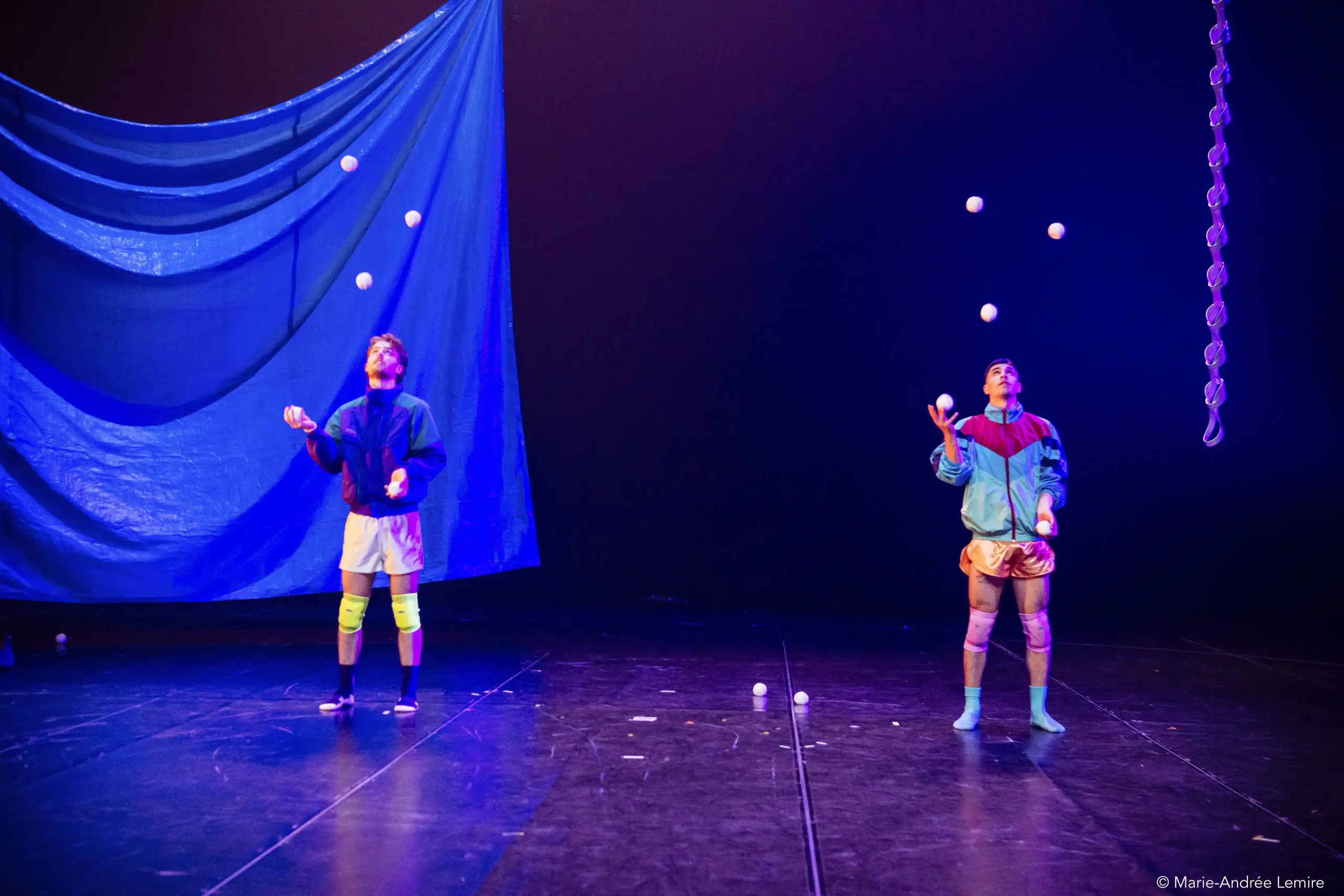Deux jongleurs, Zachary Martel et Santiago Rivera Laugerud, se produisent sur scène, chacun lançant plusieurs balles blanches en l'air. Ils portent des tenues colorées et se tiennent sous un éclairage bleu spectaculaire, sur une toile de fond bleue drapée.