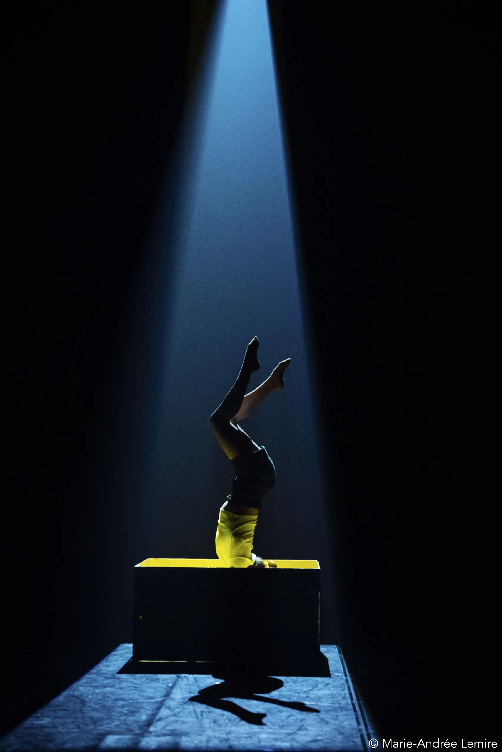 Emma Verhaeghe se tient en équilibre sur la tête au sommet d'une plate-forme jaune, éclairée par un projecteur dramatique sur un fond sombre.