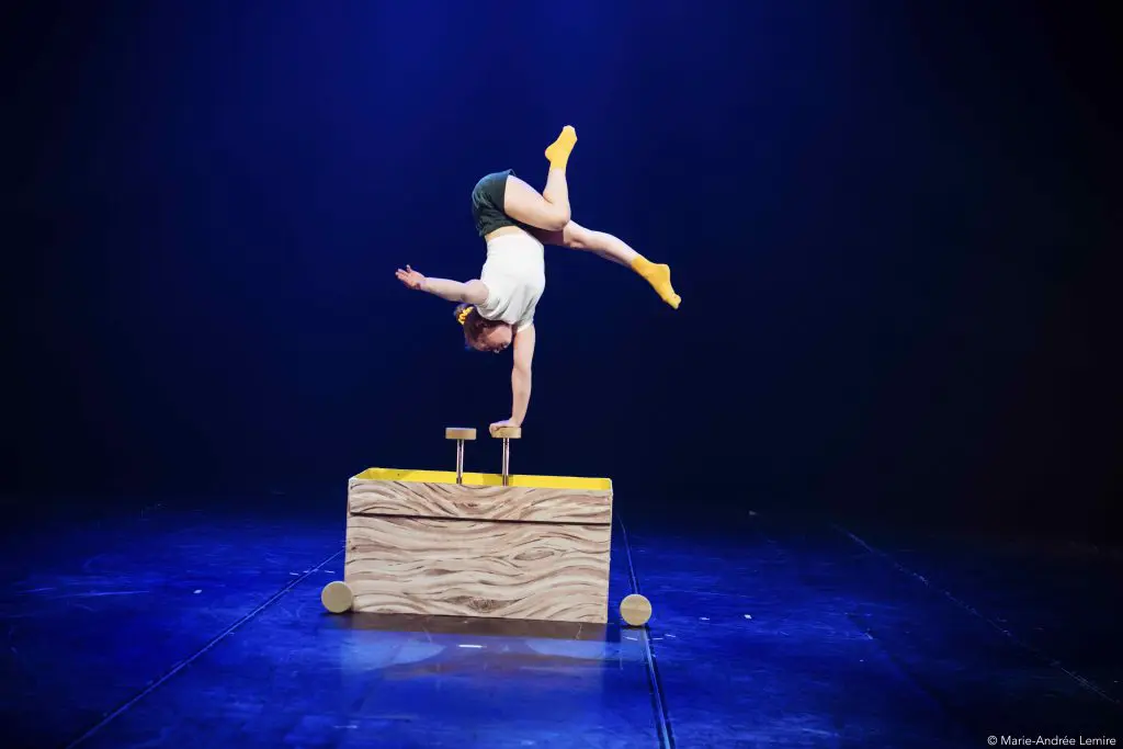Emma Verhaeghe effectue un équilibre sur une main sur un appareil de gymnastique en forme de bloc de bois, sur une scène éclairée en bleu foncé. Elle porte une chemise blanche, un short foncé et des chaussettes jaunes.