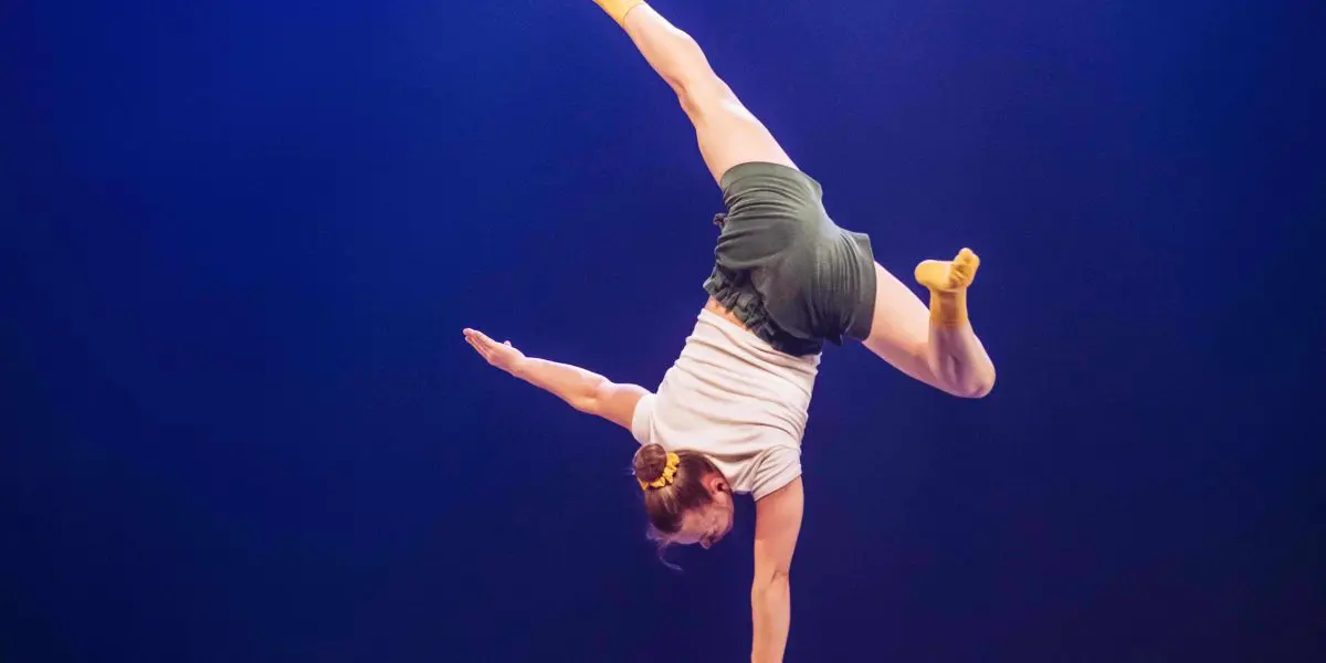 Emma Verhaeghe réalise un équilibre à une main sur une petite plate-forme avec deux supports verticaux, en se balançant dans une fente sous un éclairage de scène dramatique.