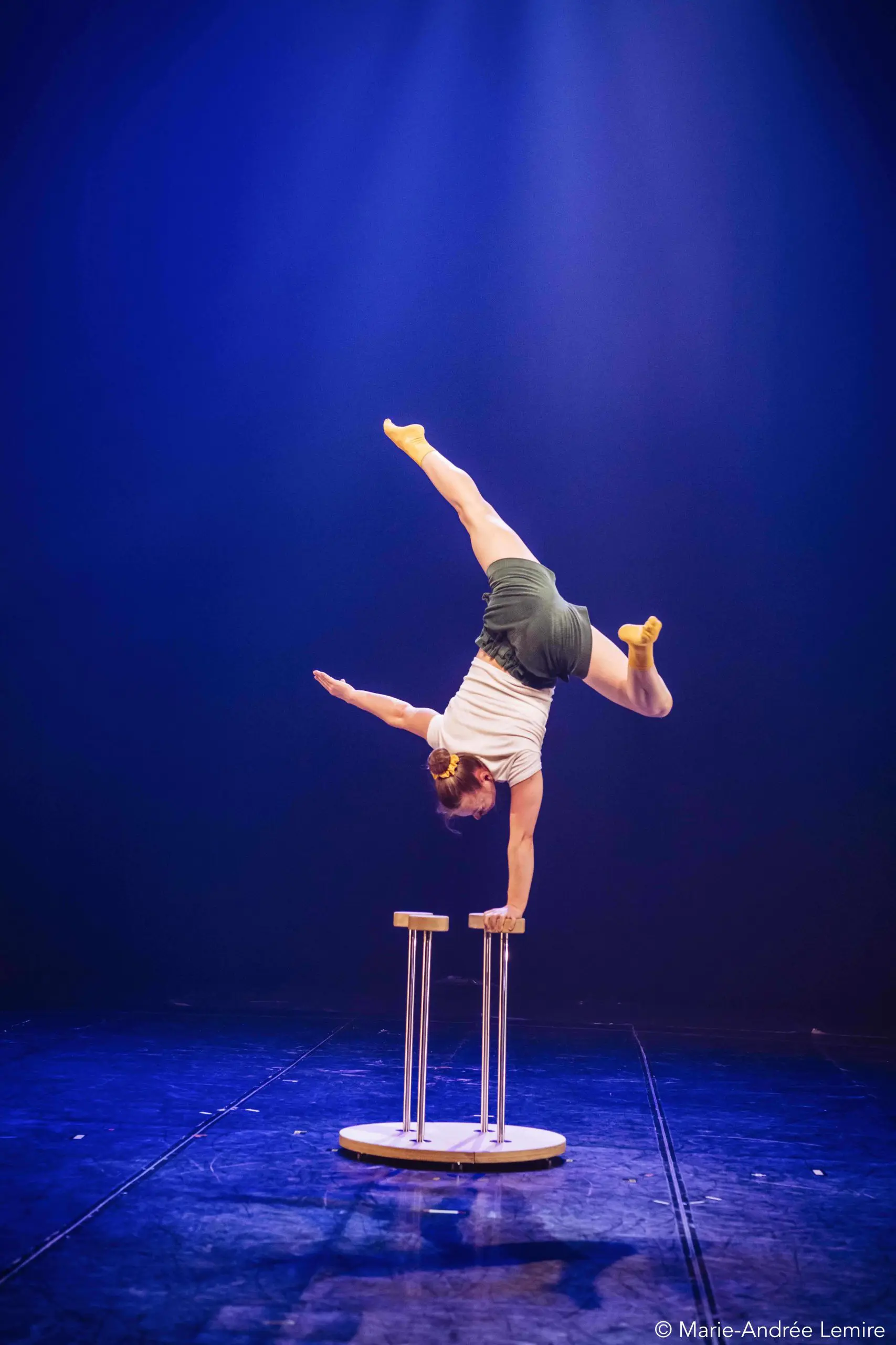 Emma Verhaeghe réalise un équilibre à une main sur une petite plate-forme avec deux supports verticaux, en se balançant dans une fente sous un éclairage de scène dramatique.
