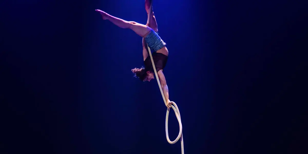 Ellie Rossi, voltigeuse émérite, exécute une pose acrobatique à l'envers sur un cerceau aérien suspendu, une jambe tendue vers le haut et l'autre accrochée, sous un éclairage de scène dramatique et sur un fond sombre.