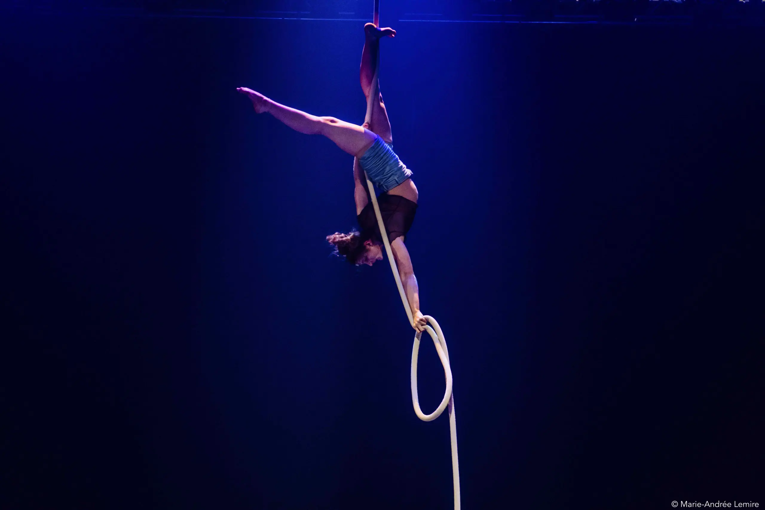 Ellie Rossi, voltigeuse émérite, exécute une pose acrobatique à l'envers sur un cerceau aérien suspendu, une jambe tendue vers le haut et l'autre accrochée, sous un éclairage de scène dramatique et sur un fond sombre.