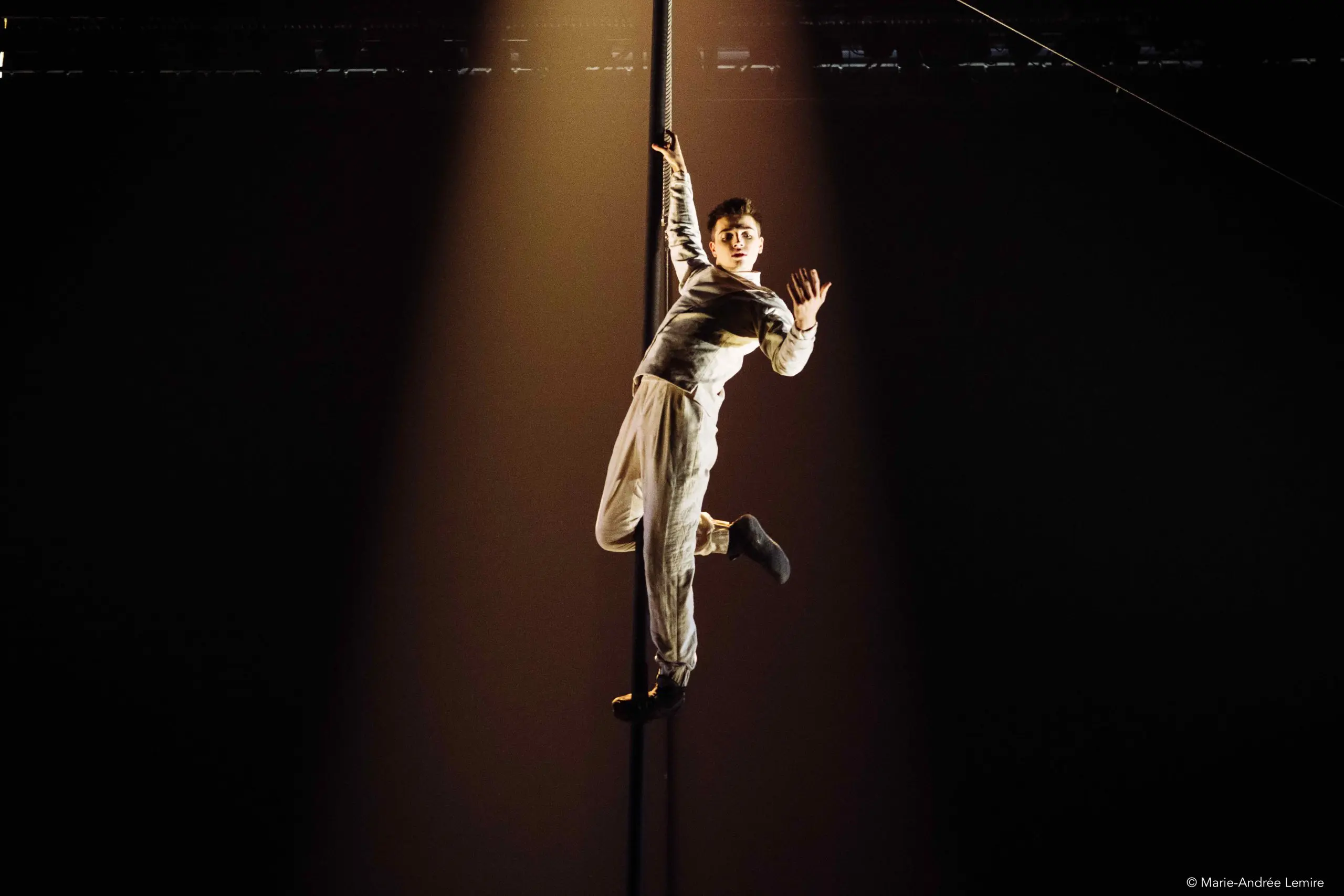 Un artiste en tenue blanche est gracieusement suspendu à un poteau vertical, éclairé par un projecteur dramatique sur un fond sombre.