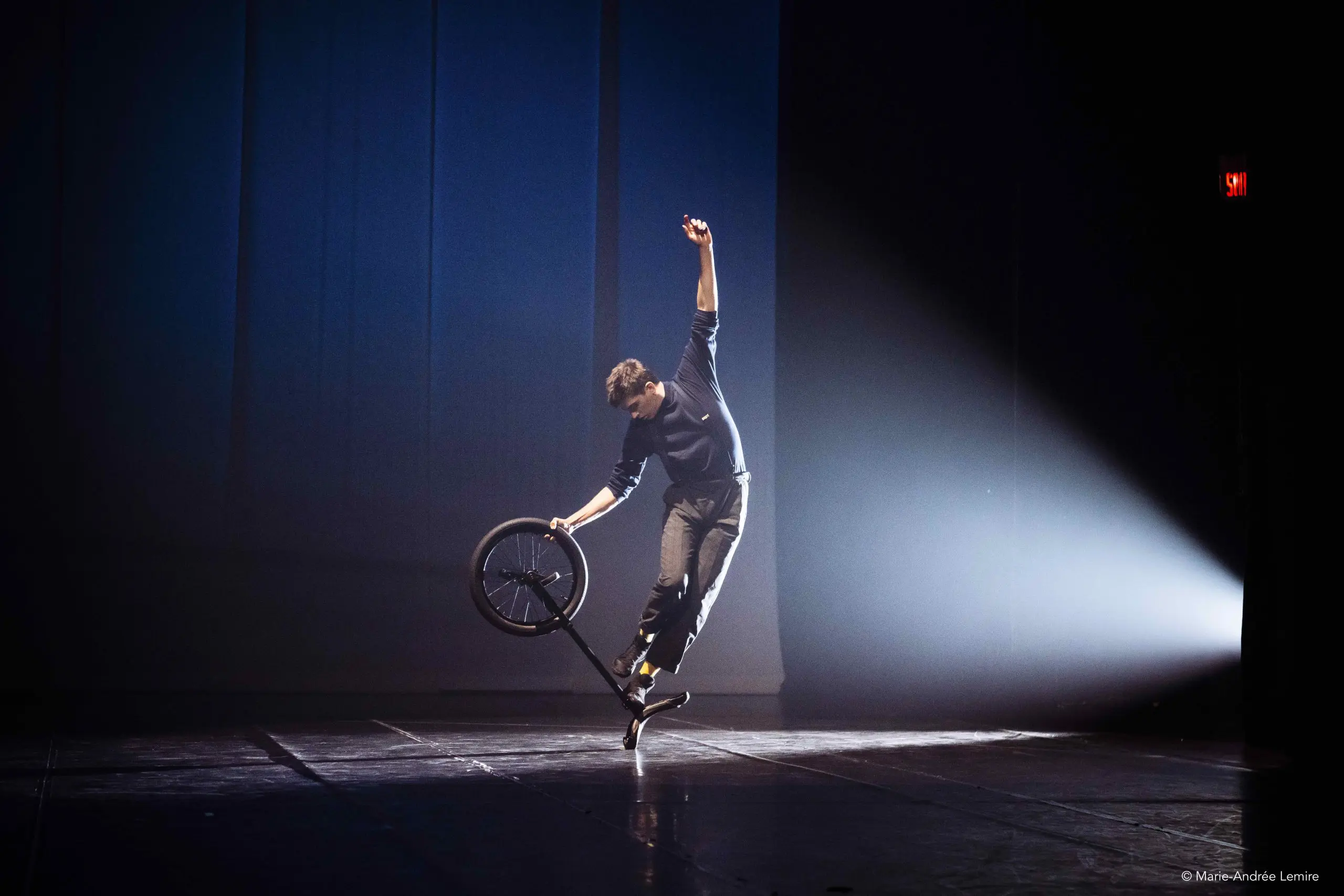 Une personne se tient en équilibre sur un pied tout en effectuant une figure avec un vélo BMX sur une scène faiblement éclairée par un seul projecteur placé sur le côté.