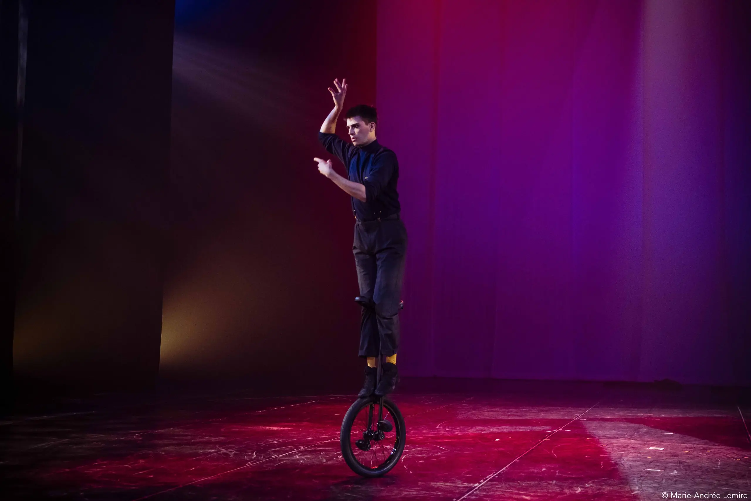 Un artiste vêtu de noir se tient en équilibre sur un monocycle sur scène, un bras levé et l'autre plié, sous un éclairage coloré rose et violet.